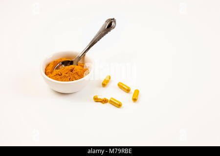 La curcuma in polvere in piccole il piatto con il cucchiaio d'argento. Curcuma capsule sulla superficie bianca. Foto Stock