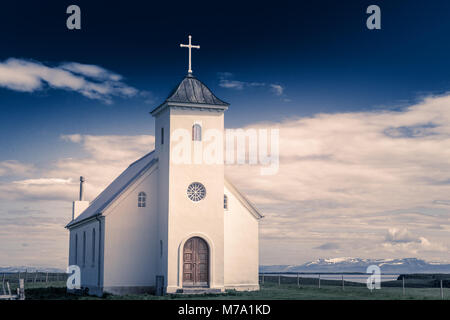 Flateyjarkirkja bianco chiesa luterana con prato in primo piano e il fiordo di mare con il cielo blu scuro e le montagne sullo sfondo, Flatey, Islanda Foto Stock