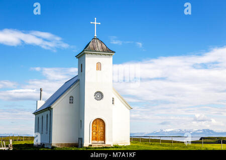 Flateyjarkirkja bianco chiesa luterana con prato in primo piano e il fiordo di mare con il blu del cielo e le montagne sullo sfondo, Flatey, Islanda Foto Stock