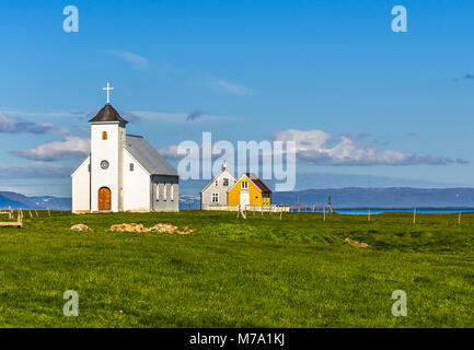 Flateyjarkirkja bianco chiesa luterana e matura della vita di capanne con prato in primo piano e il fiordo di mare con cielo blu in background, Flatey, Icel Foto Stock