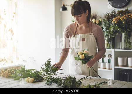 Giovani femmine fioraio mettendo insieme un mazzo di rose mentre si lavora a un tavolo nel suo negozio di fiori Foto Stock