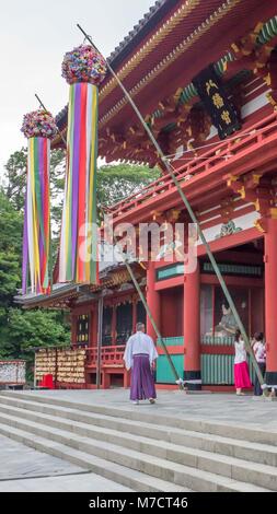 Lo Shintoismo sacerdote vestito di bianco top e pantaloni viola all ingresso della bella rossa Tsurugaoka Hachimangū santuario con grandi decorazioni colorate. Foto Stock