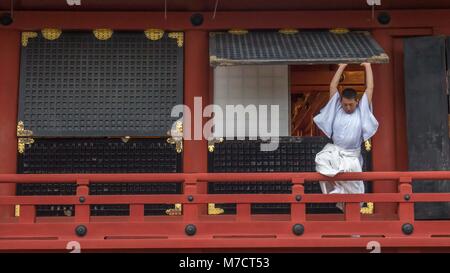 Lo Shintoismo sacerdote vestito di vesti bianche permanente sulla Tsurugaoka Hachimangū santuario della ringhiera la chiusura di grandi finestre in legno in preparazione della chiusura. Foto Stock