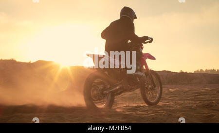 Professional FMX Motociclista torsioni pieno impugnatura a farfalla e inizia a correre sulla spiaggia sabbiosa di Off-Road via. Scenic tramonto. Foto Stock