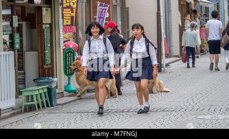 Due giovani scuola giapponese le ragazze che indossano blu e bianca uniforme sorridente e tenendo le mani a piedi a casa da scuola su una tipica strada dello shopping. Foto Stock