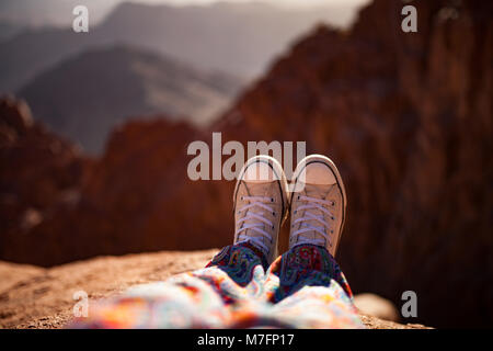 Gambe femmina in bianco gumshoes illuminata dal sole sulla scogliera contro sfondo della gamma della montagna. Foto Stock