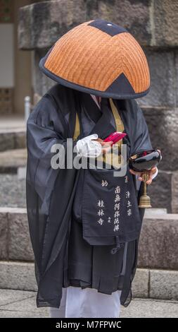 Buddista Giapponese pellegrino vestito di nero e vesti bianche e il cappello di paglia di contenimento laccato nero ciotola controllando il suo rosso incassato telefono cellulare per i messaggi. Foto Stock