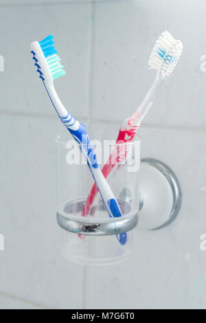 Di lui e di lei gli spazzolini da denti colorati nel becher in plastica supporto sulla parete piastrelle in bagno. Usato e segni di dentifricio sulle spazzole e bicchiere. Rosso Blu. Foto Stock