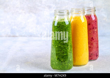 Verde, giallo, viola i frullati in bottiglie di bacche, verdi, arance su una lavagna bianca su una tabella di colore grigio Foto Stock