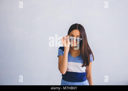 Latino la donna che indossa abiti casual guardando attraverso gli occhiali da sole e sorridente, mentre in piedi contro lo sfondo con copia spazio. Gioventù positivo Foto Stock