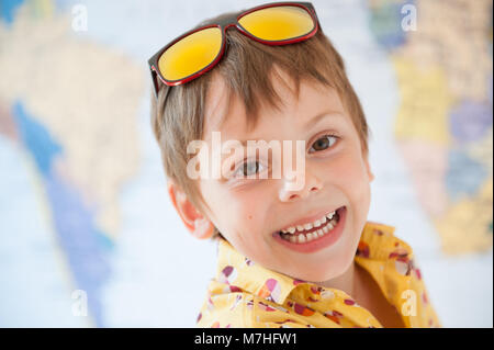 Funny sorridenti carino piccolo ragazzo in maglietta gialla e occhiali da sole sulla mappa del mondo sullo sfondo Foto Stock