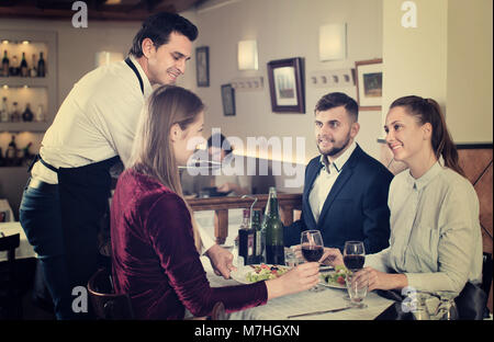 Cameriere positivo che serve pasti al cordiale compagnia gli ospiti presso il ristorante Foto Stock