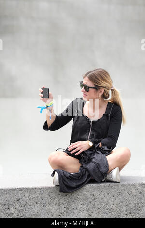 Bionda e giovane signora con la coda di cavallo, seduto sul muro con il telefono cellulare, prendendo un selfie, Bilbao Vizcaya, Pais Vasco, Spagna, Foto Stock