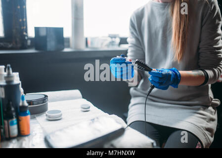 Tattooer femmina in blu guanti sterili prepara il dermografo, professional strumenti di lavoro in background. Tatuaggio in salone Foto Stock