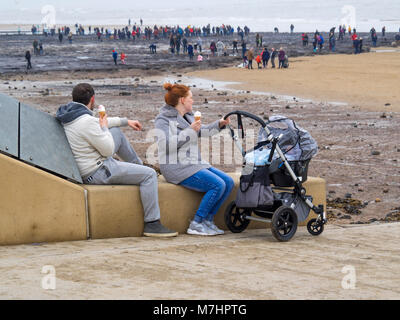 A Redcar beach una giovane coppia con un bimbo nel passeggino gustare gelati mentre la folla di gente sulla spiaggia a guardare i resti di una foresta di fossili Foto Stock