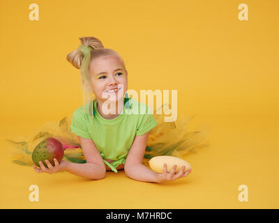 Il biondo bambina indossa una luce verde t-shirt e un luminoso mantello colorato.Tenendo il bambino tiene il mango frutti di diverse varietà. Foto Stock