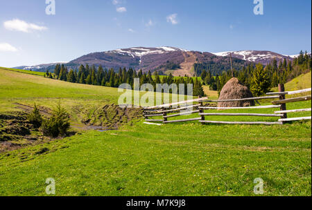 Bellissimo paesaggio rurale in primavera. staccionata in legno e pagliaio su un pendio erboso ai piedi della montagna Borzhava cresta con cime innevate. Foto Stock