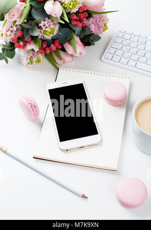 Signora blogger scrivania con fiori di colore rosa e macaron torte bianco su sfondo tabella, femminile home office workspce con caffè e lo smartphone mock-u Foto Stock