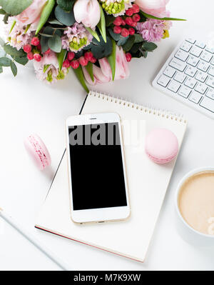 Signora blogger scrivania con fiori di colore rosa e macaron torte bianco su sfondo tabella, femminile home office workspce con caffè e lo smartphone mock-u Foto Stock