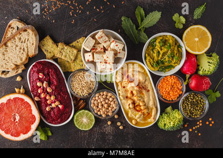 Cibo Vegan sfondo. Spuntini vegetariani: hummus, barbabietole hummus, piselli verdi dip, verdure, tofu. Vista superiore, sfondo scuro, copia dello spazio. Foto Stock