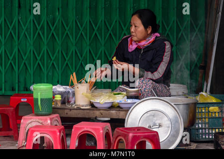 Una donna attende i clienti al suo impromptu street food in tinta, Vietnam. Le donne portano tutto ciò di cui hanno bisogno su scooter o utilizzando bambù stanghe includono un fornello, bocce, ingredienti, acqua, stock e anche le sedie in plastica. Essi impostare ovunque si può trovare uno spazio sul sentiero. Foto Stock