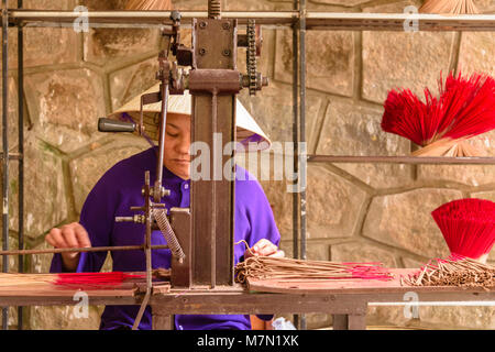 Un vietnamita donna che indossa un tradizionale di bambù cappello conico rende i bastoncini di incenso usando una vecchia macchina. Foto Stock