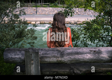 Giovane donna si siede su una panchina vicino a un fiume di montagna nel parco nel villaggio austriaco nelle Alpi. Vista posteriore di elegante ragazza nel rivestimento luminoso su in montagna. Foto Stock