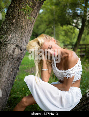 Giovane donna si siede sotto un albero nel giardino e rilassa. Malinconici ragazza bionda con gli occhi chiusi è seduto in posizione di parcheggio e sognare. Stanco di appoggio femmina Foto Stock