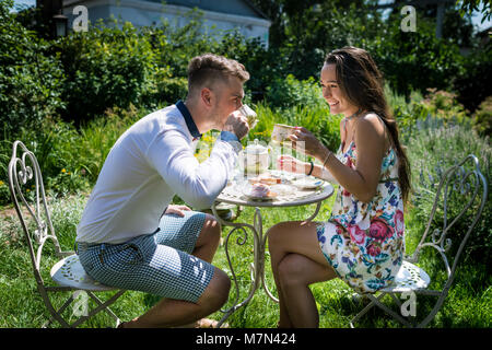 Giovane uomo si siede con la fidanzata e bevande tè con dolci. Due persone interessanti sorrisi e trattiene tazze in giardino verde. Bellissima ragazza con fidanzato Foto Stock