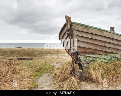 Abbandonate la barca disastrate bloccati nella sabbia. Vecchia barca in legno sulla riva sabbiosa della spiaggia. Tramonto sulla spiaggia. Vegetazione rada. Foto Stock