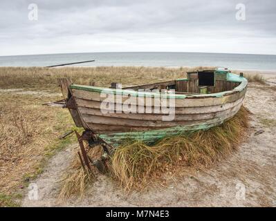 Abbandonate la barca disastrate bloccati nella sabbia. Vecchia barca in legno sulla riva sabbiosa della spiaggia. Tramonto sulla spiaggia. Vegetazione rada. Foto Stock