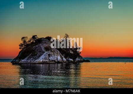 Isola rocciosa con silhouette nera di alberi e di abeti nell'oceano al tramonto della giornata contro lo sfondo delle montagne e crimson emerald sun Foto Stock