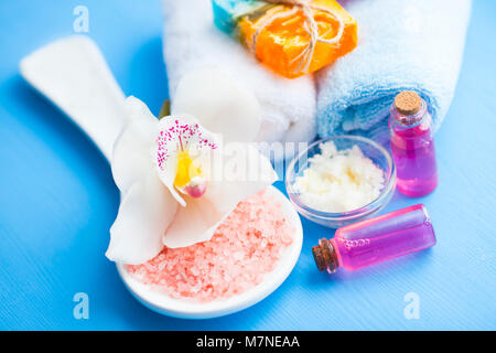 Spa e cosmetici naturali nozione. Set di pelle e cura del corpo strumenti: olio di karité, bottiglie di vetro con olio, sale marino, sapone, argilla, asciugamani, orchidea sull'azzurro Foto Stock