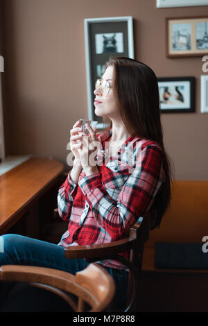 La bruna ragazza con gli occhiali e una maglietta, nel pomeriggio in un cafe dalla finestra, bevande di tè del mattino. Contiene una tazza gustando un drink. Foto Stock