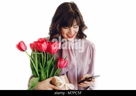 Felice madri o womens giorno concetto. Felice elegante ragazza con rosa tulipani e confezione regalo con nastro guardando al telefono e sorridente su sfondo bianco. yo Foto Stock