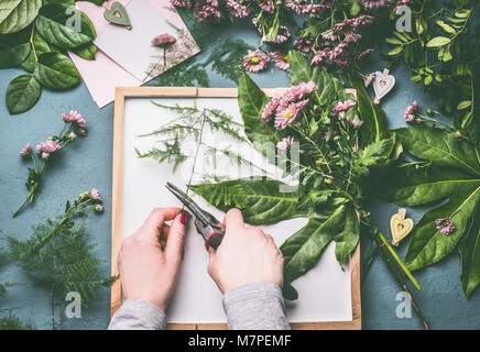 Fiorista femmina mani con le forbici facendo composizioni floreali fiori e foglie verdi sul vassoio bianco , vista superiore Foto Stock