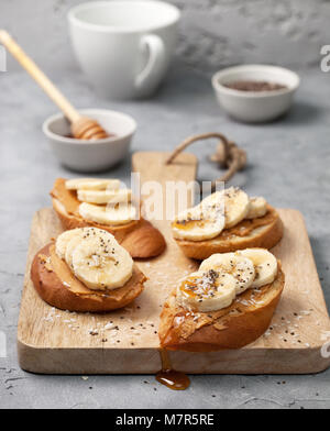 Una sana prima colazione. sandwich con burro di arachidi, banana, miele, semi di Chia e noce di cocco spruzzata su di un cemento grigio sfondo Foto Stock