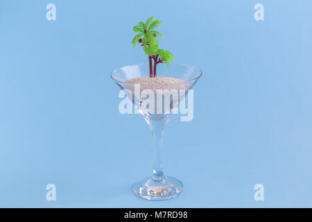Cocktail di cristallo di vetro con sabbia e palme pastello su sfondo blu. In estate la minima nozione. Foto Stock