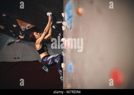 Athletic donna bouldering in una piscina centro di arrampicata. Donna scalatore holding sulle impugnature durante la salita ad una piscina palestra di arrampicata. Foto Stock