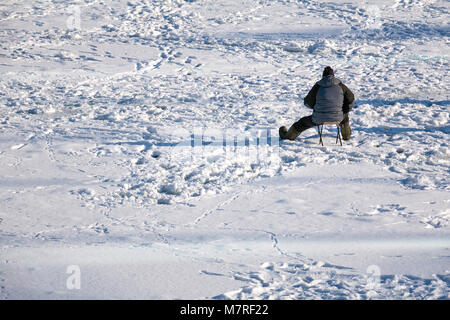 Pescatore solitario seduto in mezzo al lago ghiacciato in attesa per il pesce a comparire sulla soleggiata giornata invernale Foto Stock