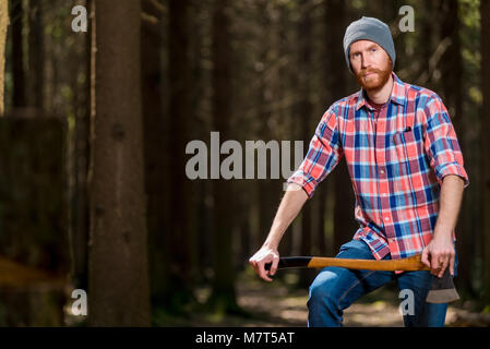 Un guardaboschi con la barba in una camicia a scacchi in posa di una foresta con un'ascia Foto Stock