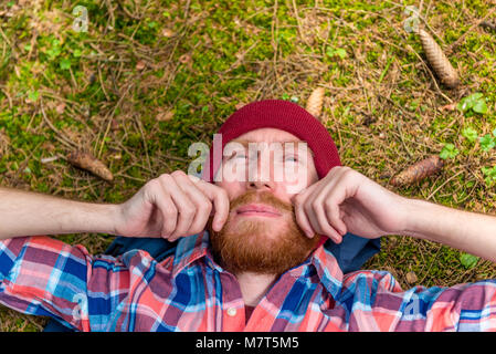 Riflessivo hipster accarezzare la sua barba rossa, un ritratto di una tenda, un uomo disteso sul terreno della foresta Foto Stock