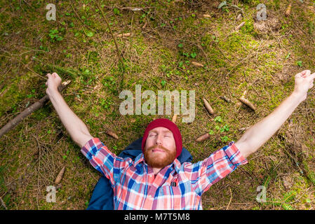 Riflessivo hipster accarezzare la sua barba rossa, un ritratto di una tenda, un uomo disteso sul terreno della foresta Foto Stock