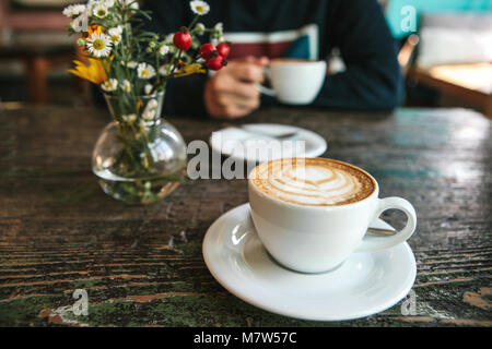 Due tazze di caffè e un vaso di fiori su un tavolo di legno, l'uomo tiene in mano una tazza di caffè in background. Una foto indica una riunione di persone e un passatempo comune. Foto Stock