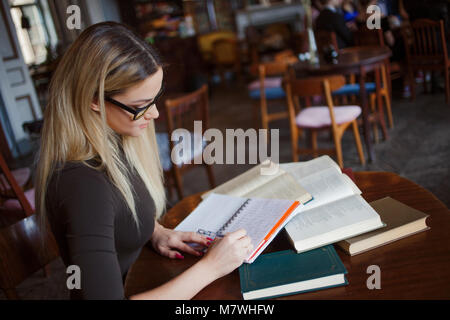 Giovane donna attraente con gli occhiali scrive in un notebook. Seduti al tavolo circondato da grande numero di libri di testo Foto Stock