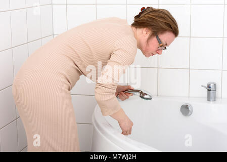 La donna caucasica, 47, con maniglia doccia per il risciacquo di una vasca da bagno nella sua casa. Indossa un abito beige e bicchieri. I suoi capelli castani è bloccata su di lei Foto Stock