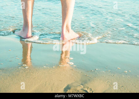 Giovane uomo caucasico maschio in piedi nudi sulla spiaggia di sabbia di mare turchese. Lavaggio onde i suoi piedi. Morbida luce del sole dorato. Colori pastello. Vacanze Rel Foto Stock