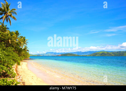 Vista della spiaggia di sabbia durante la bella giornata di sole nelle isole Fiji. Piante verdi e palme sul lato sinistro e il verde basse colline e montagne sullo sfondo. Th Foto Stock