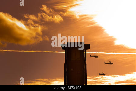 Tre elicotteri Apache volare oltre la torre con la masterizzazione di cielo arancione Foto Stock