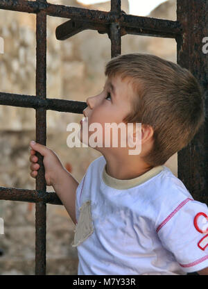 Ragazzo in piedi e guardando in alto dietro un antico cancello di ferro sul castello di greco Foto Stock